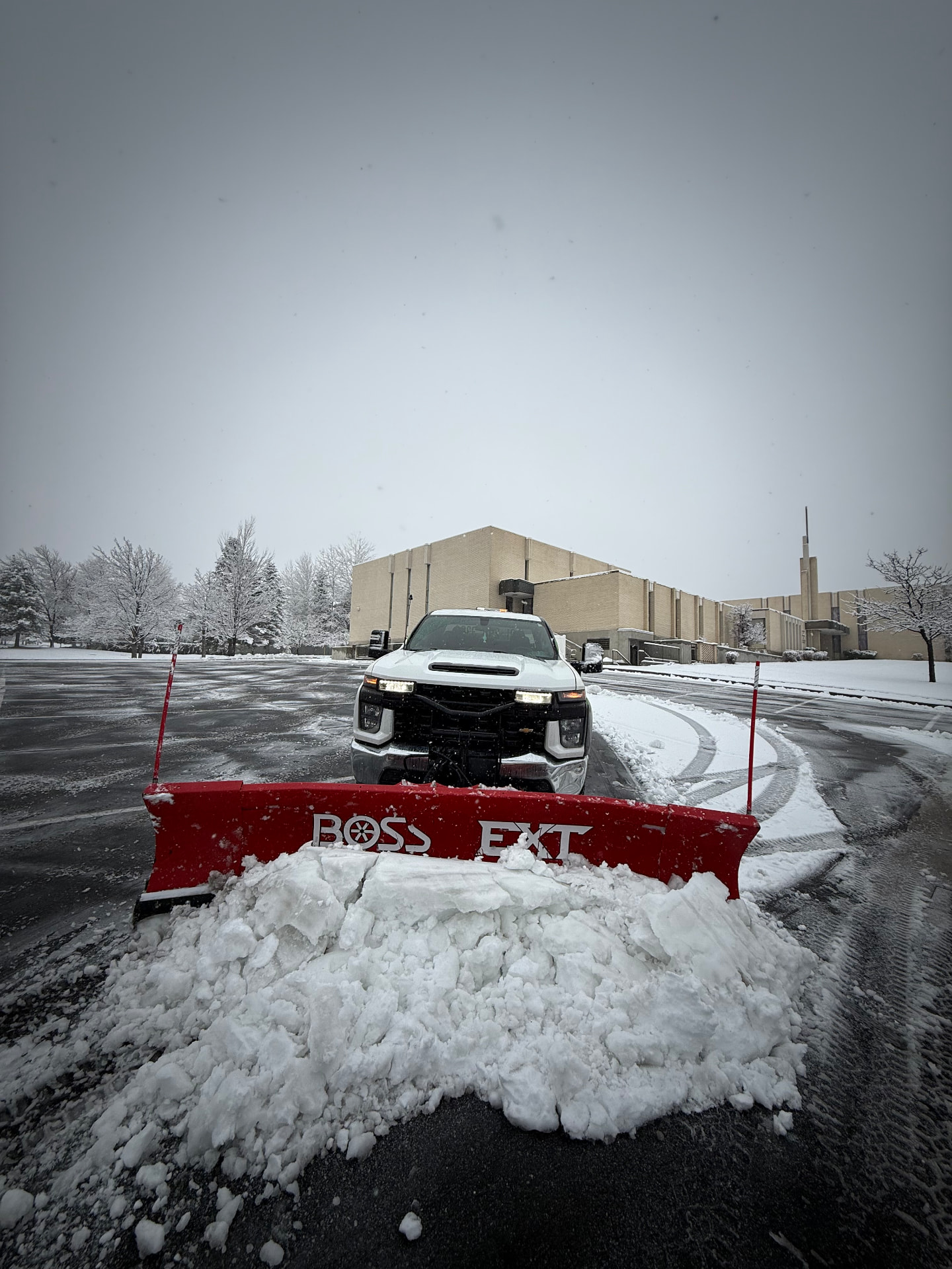 White pickup truck with a red snowplow pushing a load of snow in front of a brick building